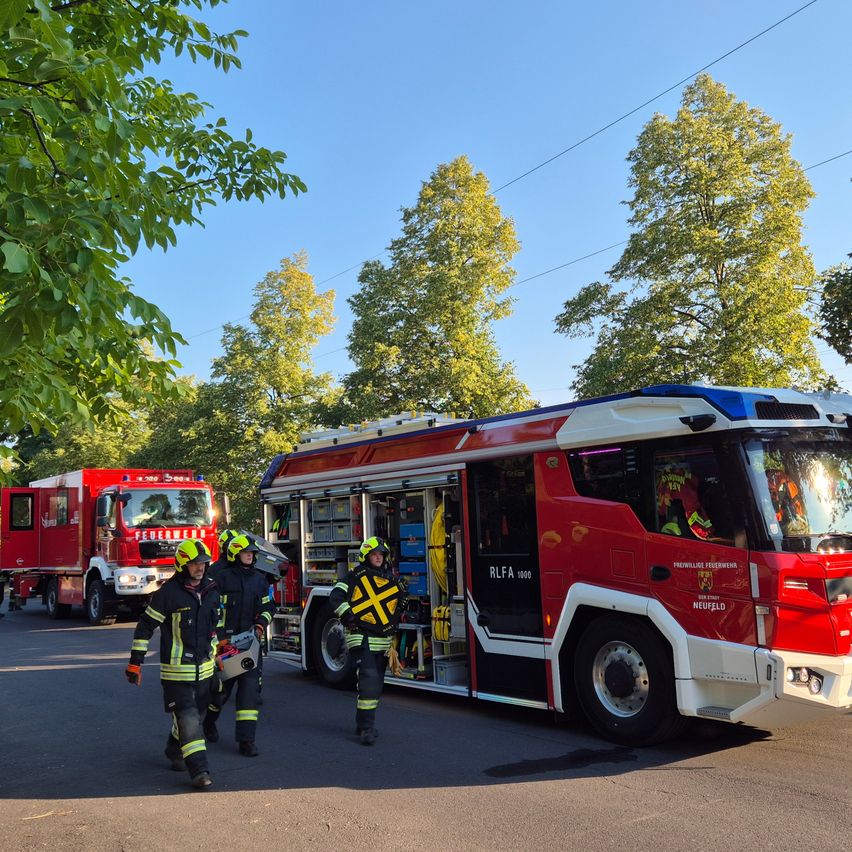 Drei Feuerwehrleute gehen auf einen roten Feuerwehrwagen mit offenen Türen zu, der am Straßenrand geparkt ist. Ein weiterer Feuerwehrwagen ist dahinter geparkt. Bäume und elektrische Leitungen sind im Hintergrund.
