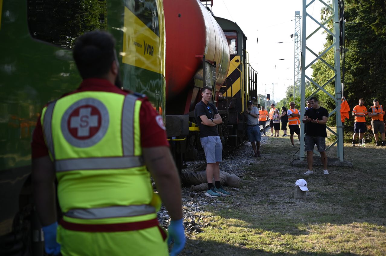 Ein Mann in einer gelben Warnweste steht neben einem Zug mit einem roten Tankwagen. Mehrere Personen in orangefarbenen Warnwesten sind auf den Gleisen versammelt, möglicherweise bei einem Vorfall.