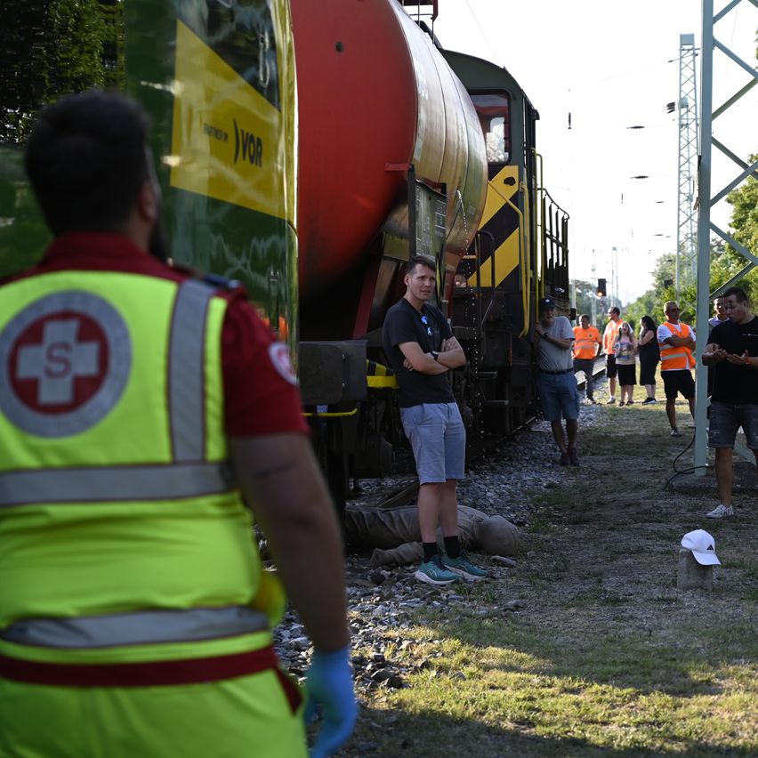 Ein Mann in einer gelben Warnweste steht neben einem Zug mit einem roten Tankwagen. Mehrere Personen in orangefarbenen Warnwesten sind auf den Gleisen versammelt, möglicherweise bei einem Vorfall.