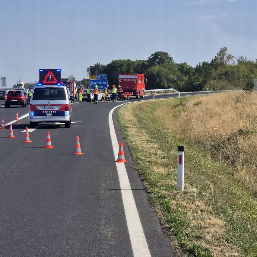 Ein Polizeiwagen mit Warnblinkanlage steht am Straßenrand, mit Verkehrskegeln, die den Bereich abgrenzen. Mehrere Personen in Sicherheitswesten kümmern sich in der Mitte der Straße um eine Situation. Ein Feuerwehrwagen und ein roter LKW sind in der Nähe geparkt.
