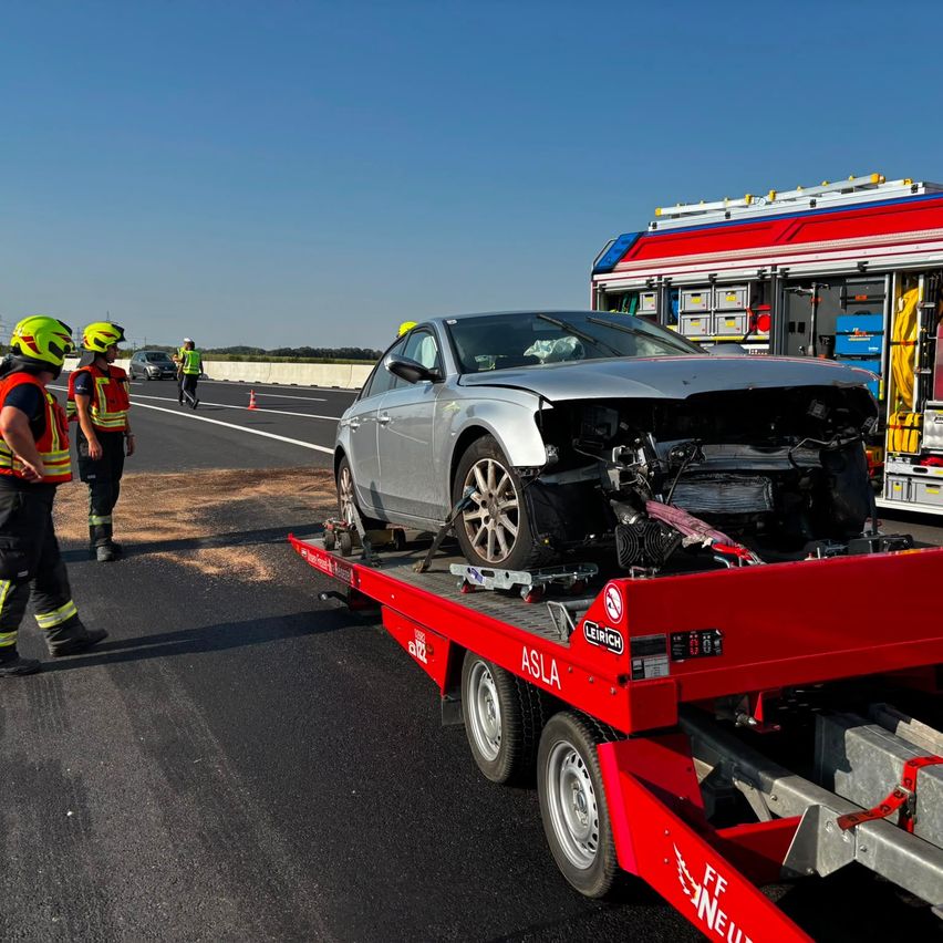 Ein zerbeultes silbernes Auto wird auf einem roten Tieflader abtransportiert. Zwei Arbeiter in Warnwesten und Helmen stehen in der Nähe. Ein Feuerwehrwagen ist am Straßenrand geparkt.