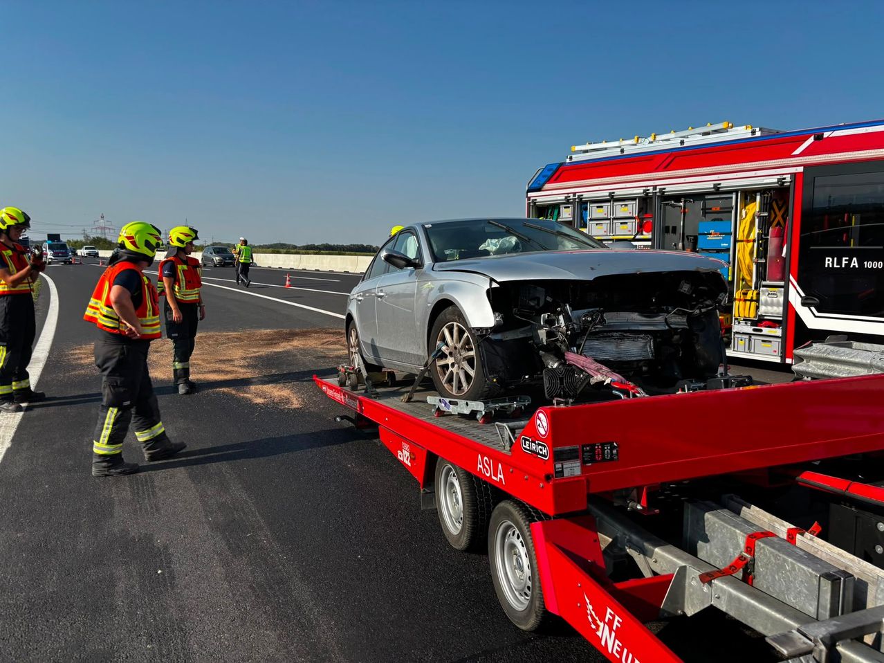 Ein zerbeultes silbernes Auto wird auf einem roten Tieflader abtransportiert. Zwei Arbeiter in Warnwesten und Helmen stehen in der Nähe. Ein Feuerwehrwagen ist am Straßenrand geparkt.