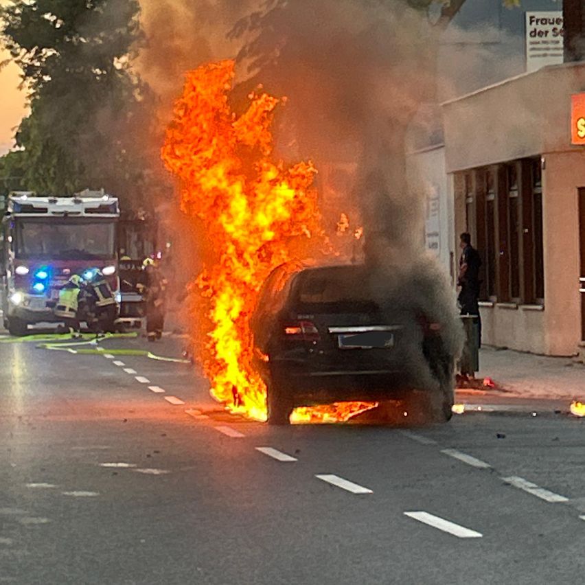 Ein Auto steht in Flammen auf der Straße, Feuerwehrleute und ein Feuerwehrwagen sind in der Nähe.