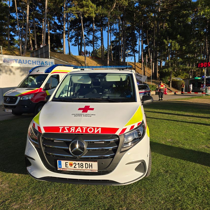 Ein Krankenwagen mit dem Wort 'Notarzt' auf der Front ist auf einem Rasenfeld geparkt, mit einem weiteren Krankenwagen und einem Gebäude im Hintergrund.