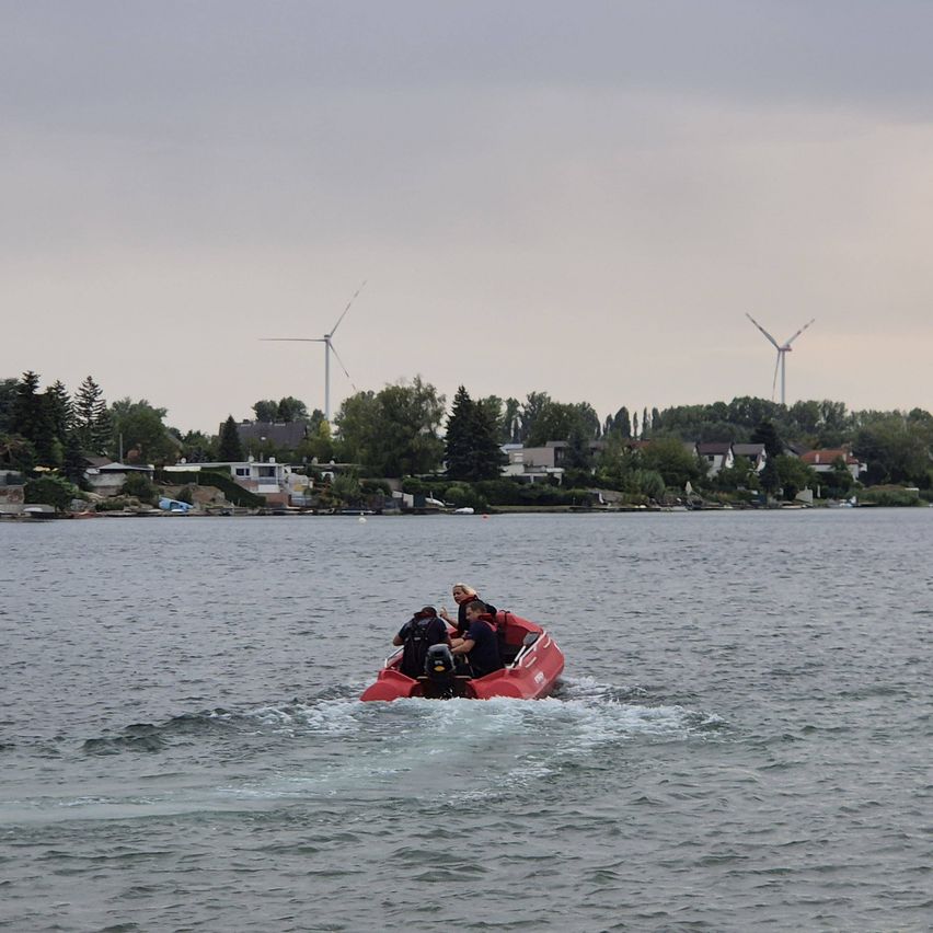 Drei Personen fahren in einem roten Boot auf einem See, wobei zwei Windturbinen in der Ferne sichtbar sind.