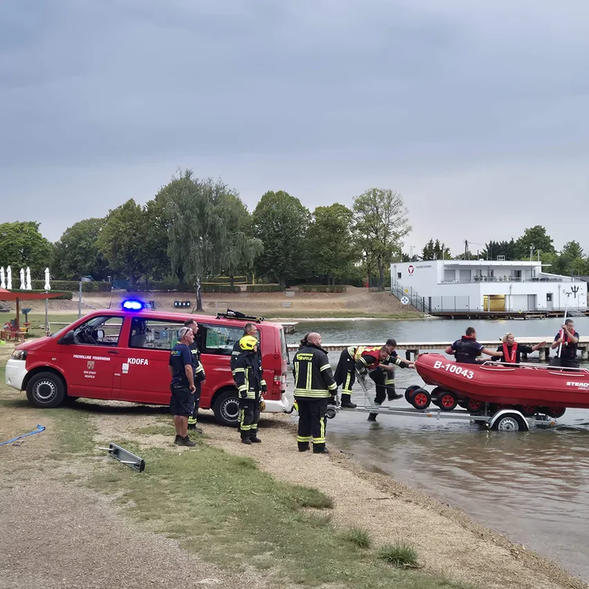 Eine Gruppe von Feuerwehrleuten in Uniform bereitet sich darauf vor, ein rotes Boot vom Ufer ins Wasser zu bringen. Das Boot steht auf einem Anhänger, und die Feuerwehrleute stehen darum herum. Ein roter Van mit eingeschalteten Notfallleuchten ist in der Nähe geparkt.