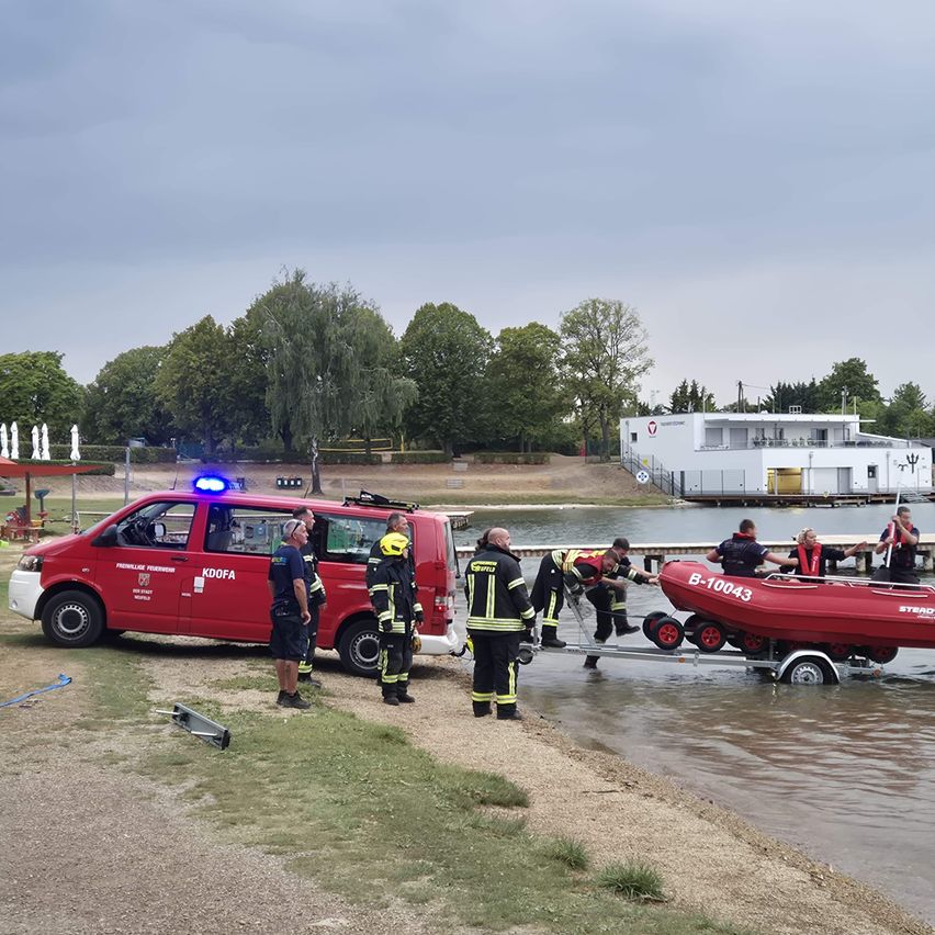 Eine Gruppe von Feuerwehrleuten in Uniform bereitet sich darauf vor, ein rotes Boot vom Ufer ins Wasser zu bringen. Das Boot steht auf einem Anhänger, und die Feuerwehrleute stehen darum herum. Ein roter Van mit eingeschalteten Notfallleuchten ist in der Nähe geparkt.