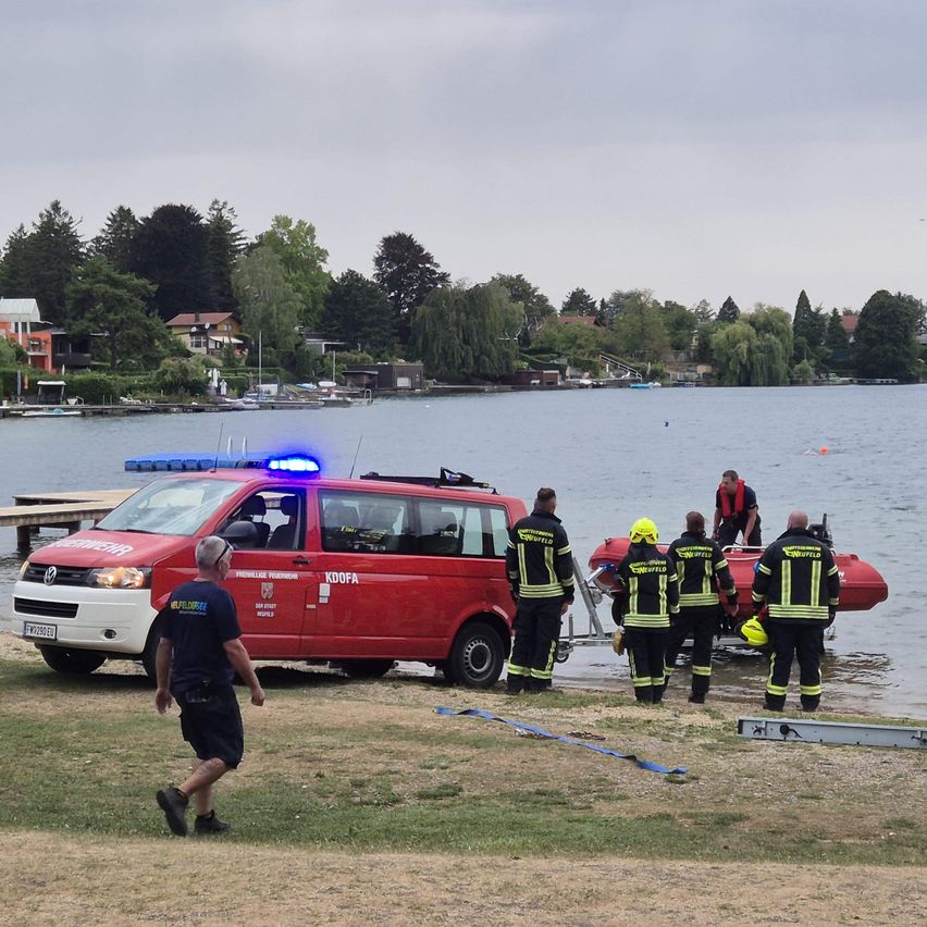 Ein rotes Notfallfahrzeug ist am Ufer geparkt, während Feuerwehrleute in gelben und schwarzen Uniformen sich darauf vorbereiten, ein rotes Boot ins Wasser zu bringen. Ein Mann in einem roten Rettungsweste steht auf dem Boot.