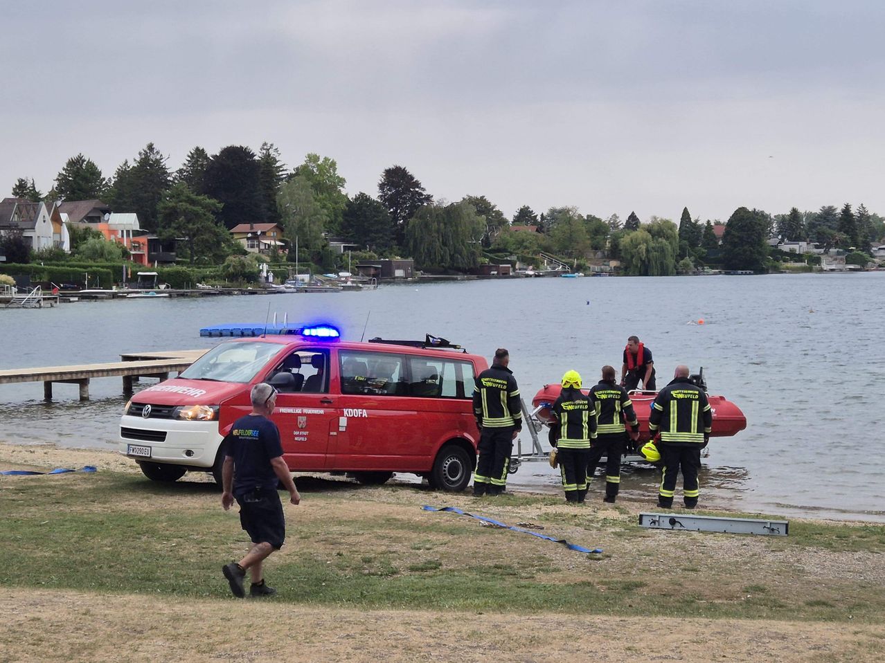 Ein rotes Notfallfahrzeug ist am Ufer geparkt, während Feuerwehrleute in gelben und schwarzen Uniformen sich darauf vorbereiten, ein rotes Boot ins Wasser zu bringen. Ein Mann in einem roten Rettungsweste steht auf dem Boot.