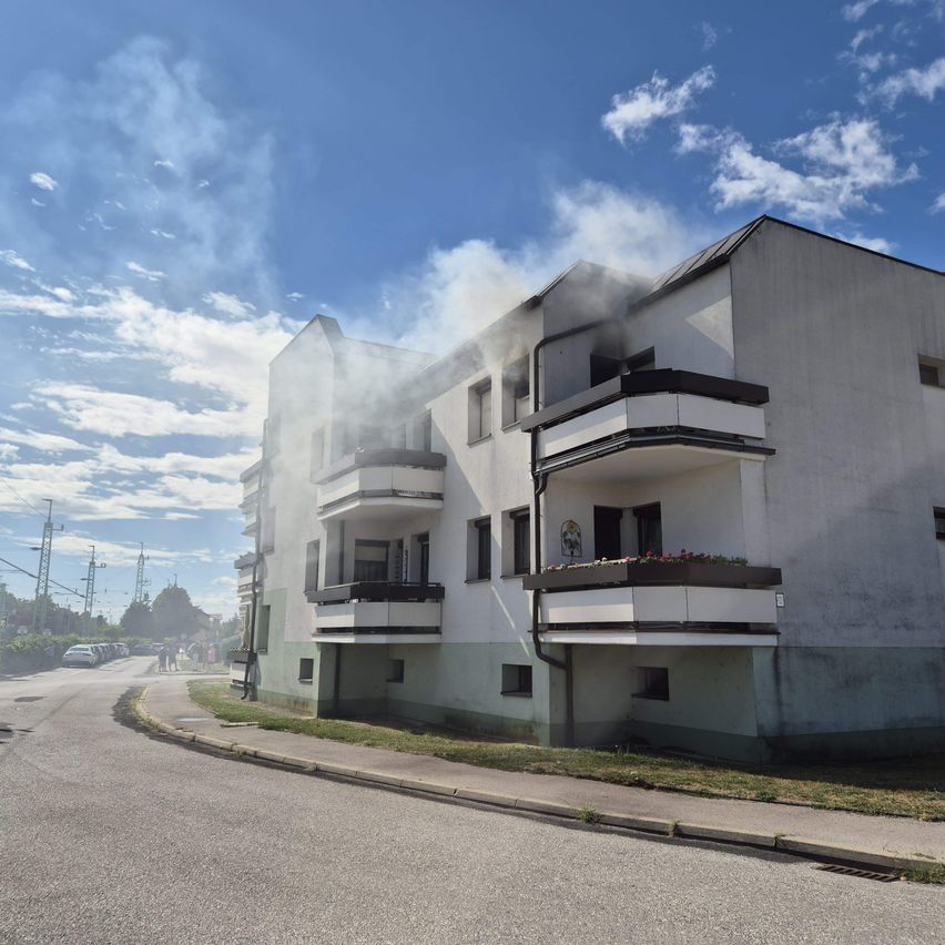 Ein mehrstöckiges Wohnhaus mit Rauch, der aus einem der oberen Stockwerke kommt. Das Gebäude hat mehrere Balkone und Autos sind am Straßenrand geparkt.