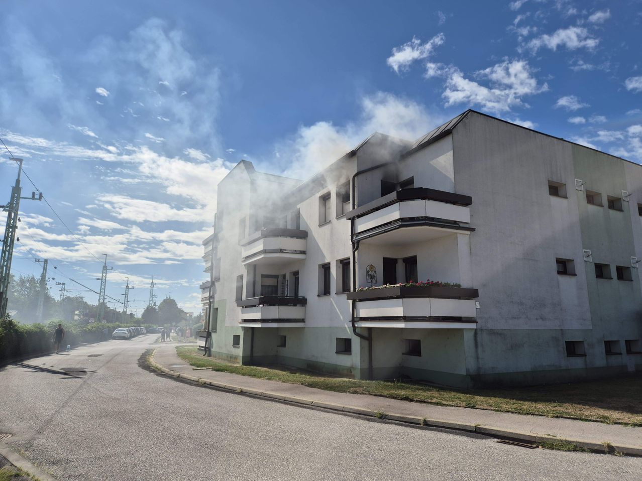 Ein mehrstöckiges Wohnhaus mit Rauch, der aus einem der oberen Stockwerke kommt. Das Gebäude hat mehrere Balkone und Autos sind am Straßenrand geparkt.