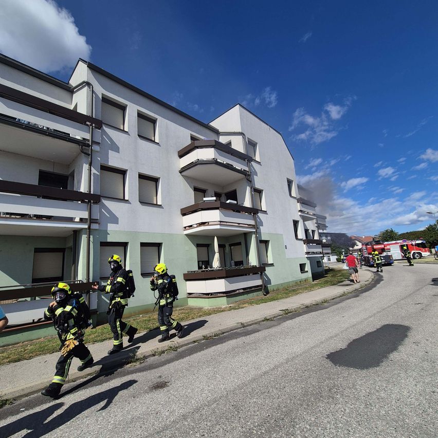 Feuerwehrleute in Schutzkleidung rennen auf ein Gebäude zu, aus dessen Balkon Rauch aufsteigt.