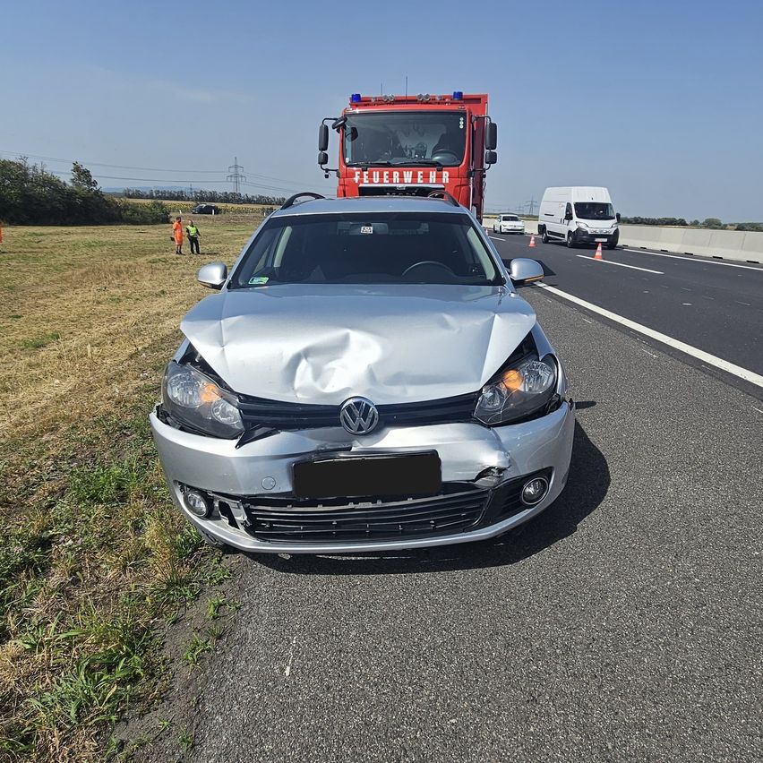 Ein silberner Volkswagen-Wagen mit beschädigtem vorderen Stoßfänger steht am Straßenrand. Im Hintergrund befinden sich ein roter Feuerwehrwagen und ein weißer Lieferwagen.