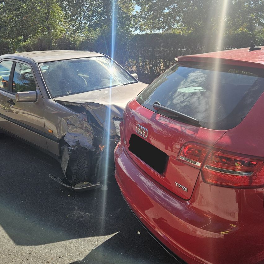 A silver car with a damaged bumper and a red car with a license plate reading TFSI. Both cars are parked on the side of the road.