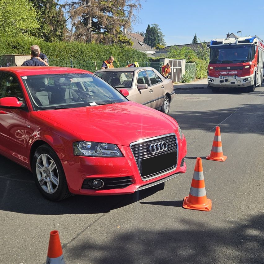 Ein roter Audi-Wagen ist auf der Straße geparkt, mit Verkehrskegel drum herum. Ein Feuerwehrwagen ist in der Nähe geparkt, und mehrere Leute stehen herum.