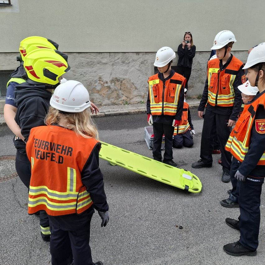Eine Gruppe von Kindern, die Feuerwehruniformen und Helme tragen, steht um eine gelbe Trage auf der Straße. Eine Frau macht ein Foto im Hintergrund.