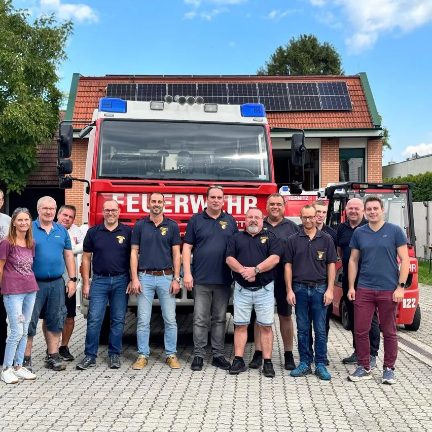 Eine Gruppe von Menschen steht vor einem Feuerwehrauto mit Solarpanelen auf dem Dach. Sie sind in Uniformen und Freizeitkleidung gekleidet. Das Feuerwehrauto ist vor einem Gebäude mit einem Ziegeldach geparkt.