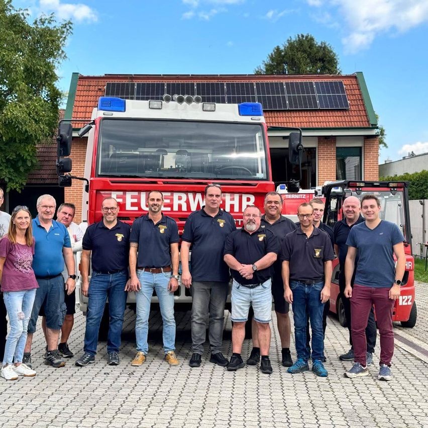 Eine Gruppe von Menschen steht vor einem Feuerwehrauto mit Solarpanelen auf dem Dach. Sie sind in Uniformen und Freizeitkleidung gekleidet. Das Feuerwehrauto ist vor einem Gebäude mit einem Ziegeldach geparkt.