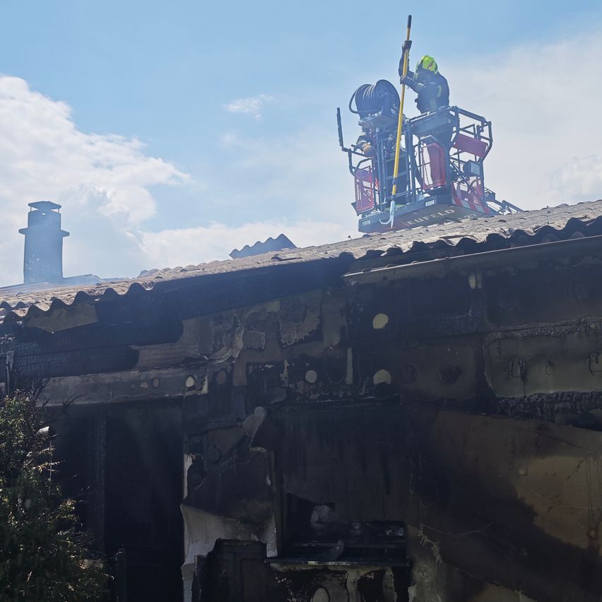 Ein Feuerwehrmann auf einer Leiter auf einem Dach mit einer Feuerwehrspritze in der Hand, neben einer anderen Person stehend. Das Gebäude ist beschädigt und verkohlt.