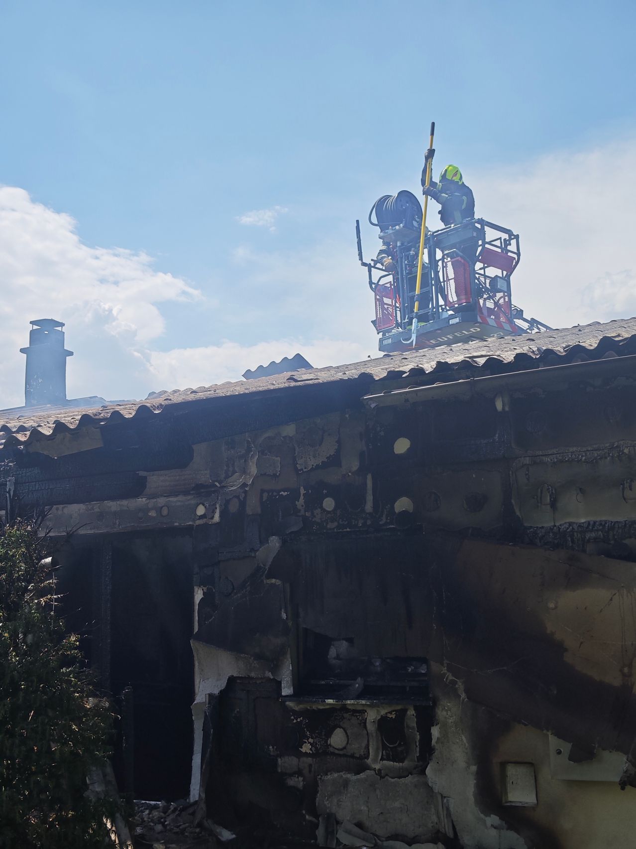 Ein Feuerwehrmann auf einer Leiter auf einem Dach mit einer Feuerwehrspritze in der Hand, neben einer anderen Person stehend. Das Gebäude ist beschädigt und verkohlt.