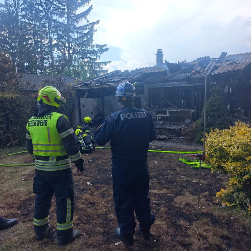 Einsatzkräfte stehen vor einem ausgebrannten Gebäude. Ein Feuerwehrmann in einem gelben Weste schaut zu, während ein Polizeibeamter in einem blauen Uniform nach unten schaut.