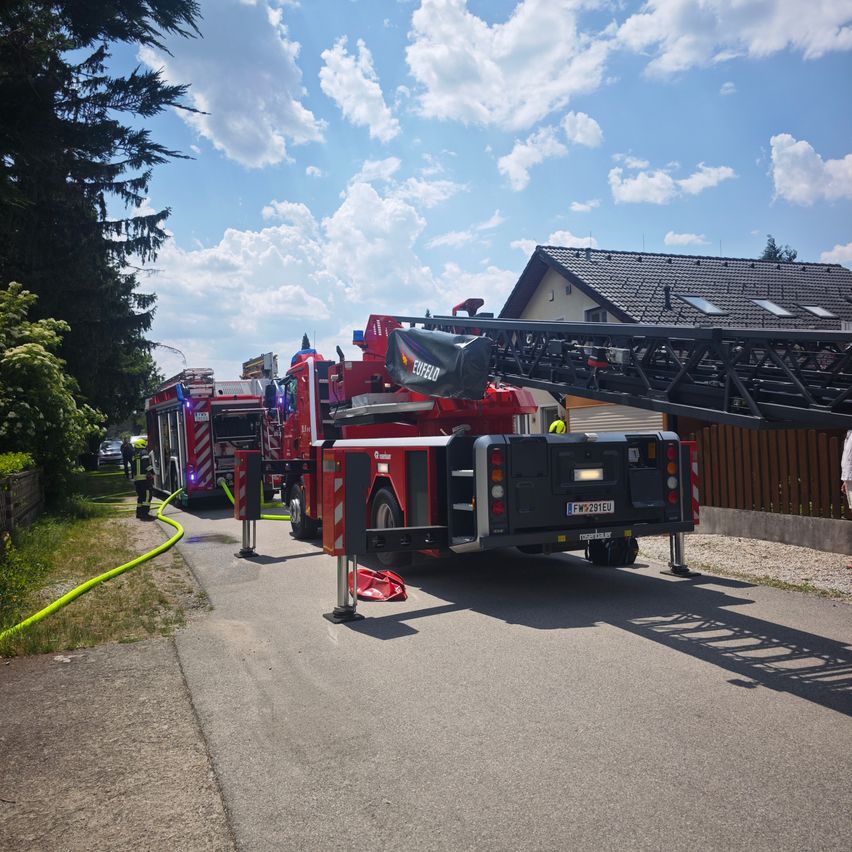 Zwei Feuerwehrfahrzeuge sind an einem sonnigen Tag in der Nähe eines Hauses geparkt. Die Leiter eines Fahrzeugs ist ausgefahren, und eine Person steht in der Nähe.