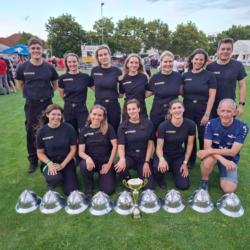 Eine Gruppe von Frauen und zwei Männern posiert mit Helmen und einem Pokal auf einem Rasenfeld.