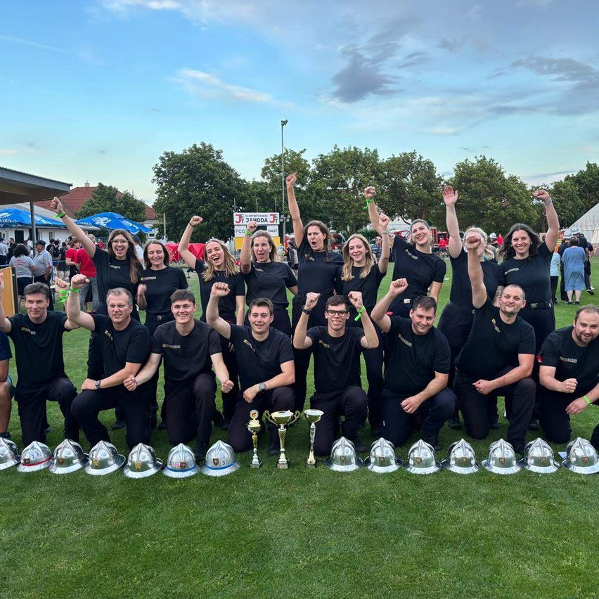 Eine Gruppe Feuerwehrleute in einheitlichen schwarzen Uniformen posiert für ein Foto mit silbernen Helmen und Trophäen, wahrscheinlich zur Feier eines Sieges bei einem Event.