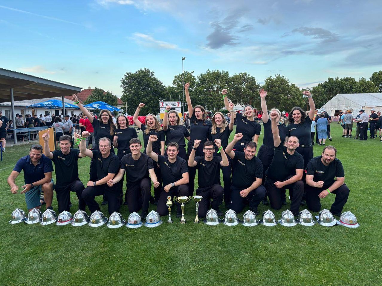 Eine Gruppe Feuerwehrleute in einheitlichen schwarzen Uniformen posiert für ein Foto mit silbernen Helmen und Trophäen, wahrscheinlich zur Feier eines Sieges bei einem Event.