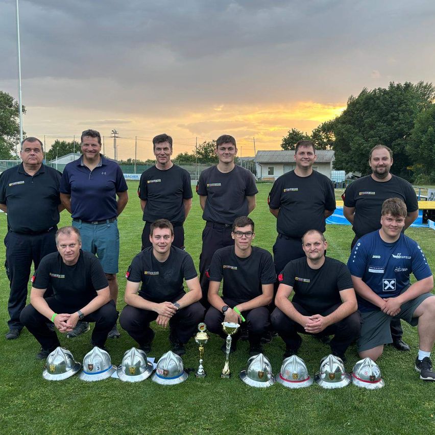 Eine Gruppe von Männern in gleichen schwarzen Uniformen posiert für ein Foto auf einem Feld mit Trophäen. Dahinter ist ein bewölkter Himmel und einige Bäume zu sehen.