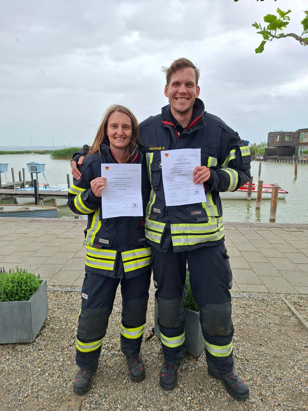 Zwei Personen in Feuerwehruniformen halten Urkunden und stehen am Wasser mit einem Steg im Hintergrund.