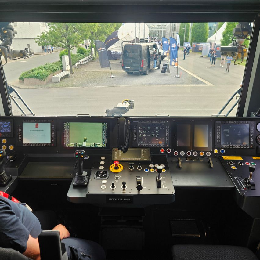 Blick aus dem Cockpit eines Fahrzeugs mit Monitoren und Steuerungen. Draußen fährt ein Lieferwagen an einem Park mit Bäumen und spazierengehenden Menschen vorbei.