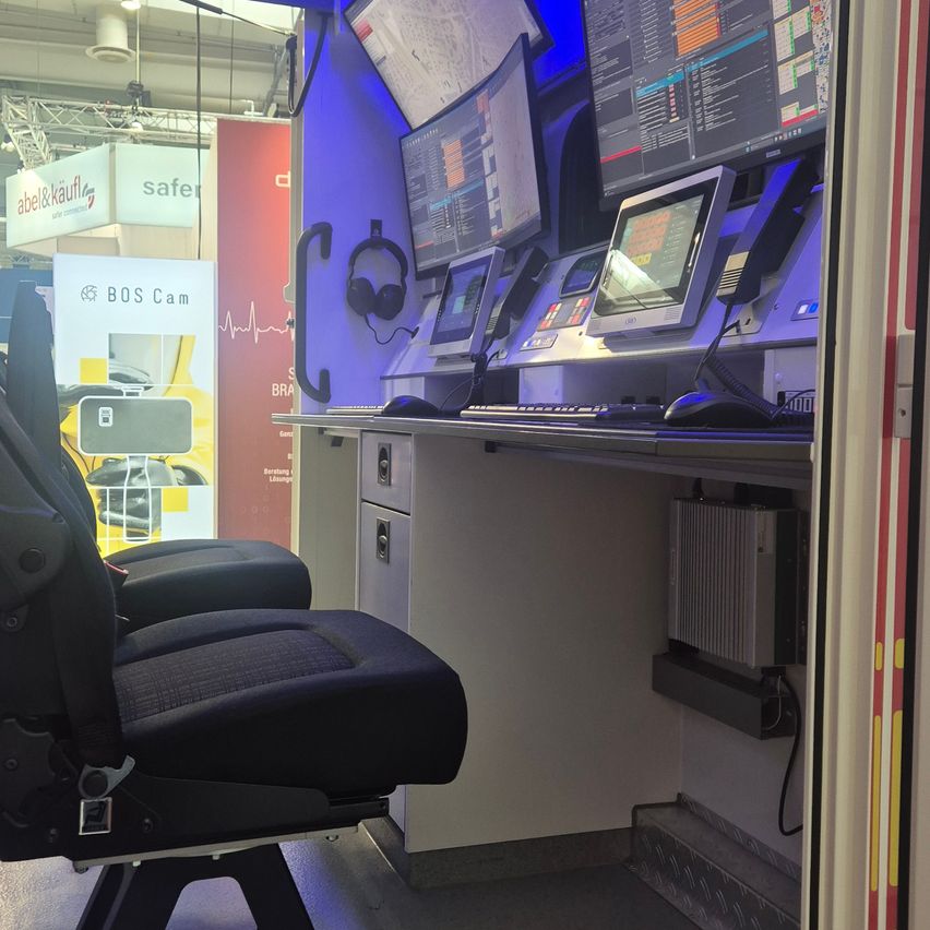 Innenraum eines Rettungswagens mit schwarzen Stühlen, mehreren Monitoren, einer Tastatur und einer Maus. Ein Schild für BOS Cam ist im Hintergrund sichtbar.