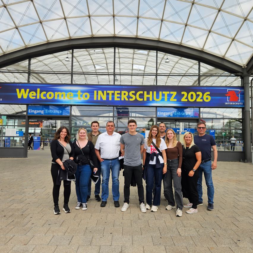 Eine Gruppe von Menschen steht vor einem großen Gebäude mit dem Schild Willkommen bei INTERSCHUTZ 2026.