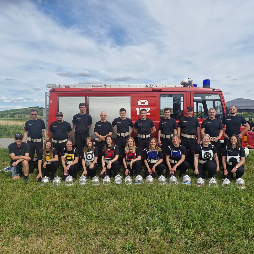 Eine Gruppe Feuerwehrleute posiert für ein Foto mit einem Feuerwehrauto, Helmen und Trophäen auf einer Wiese.