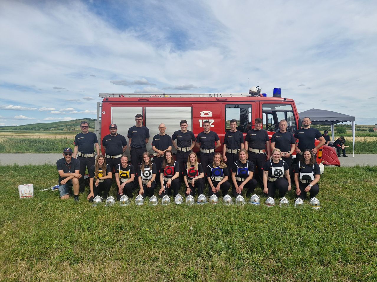 Eine Gruppe Feuerwehrleute posiert für ein Foto mit einem Feuerwehrauto, Helmen und Trophäen auf einer Wiese.