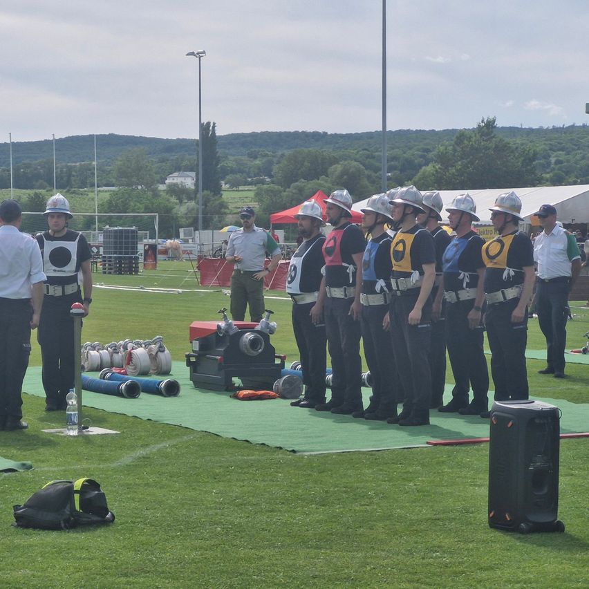 Eine Gruppe von Menschen in Uniformen steht auf einem grünen Feld. Sie halten eine Zielscheibe und scheinen sich auf eine Veranstaltung vorzubereiten.