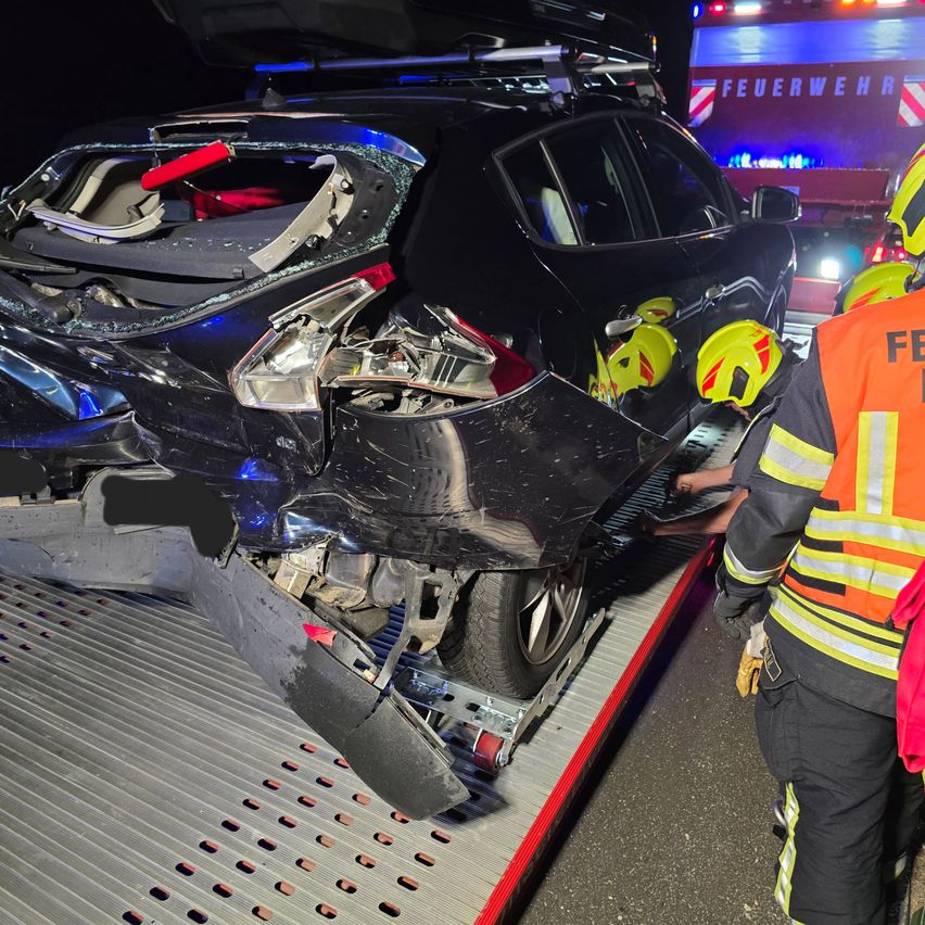 Ein stark beschädigtes schwarzes Auto wird von Feuerwehrleuten abgeschleppt. Die Front ist zerdrückt und die Motorhaube ist offen. Im Hintergrund blinken Notleuchten.