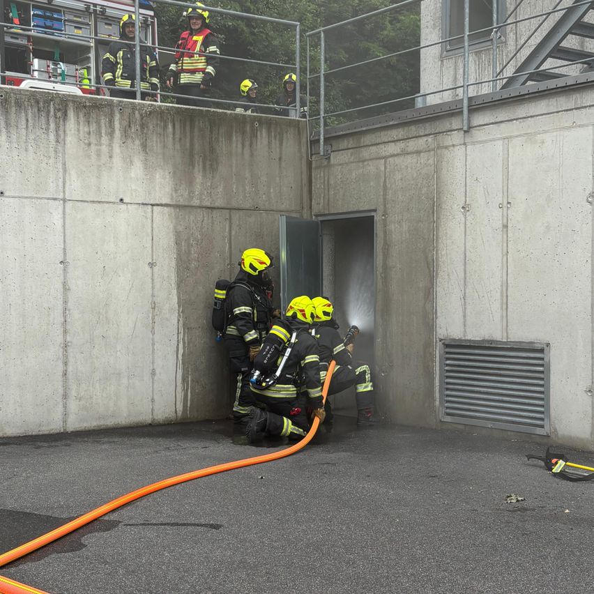 Firefighters are working to open a door in a tunnel. They are wearing yellow helmets and safety gear. Some are using a hose.