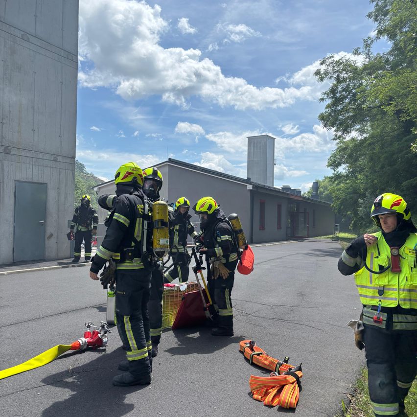 Firefighters are preparing for a training exercise on a sunny day, wearing protective gear and carrying equipment. They are positioned on a road with buildings in the background.