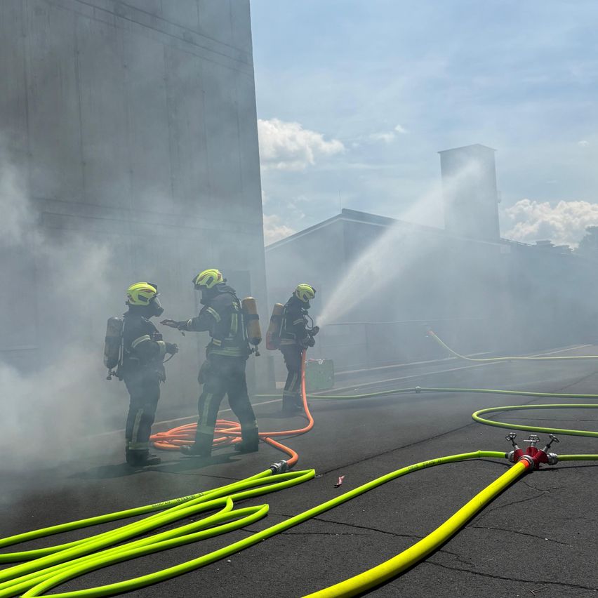 Drei Feuerwehrleute arbeiten an einem Feuer mit einer Schlauchleitung, einer sprüht Wasser, während die anderen beiden bereitstehen mit ihrer Ausrüstung in einem nebligen Bereich.