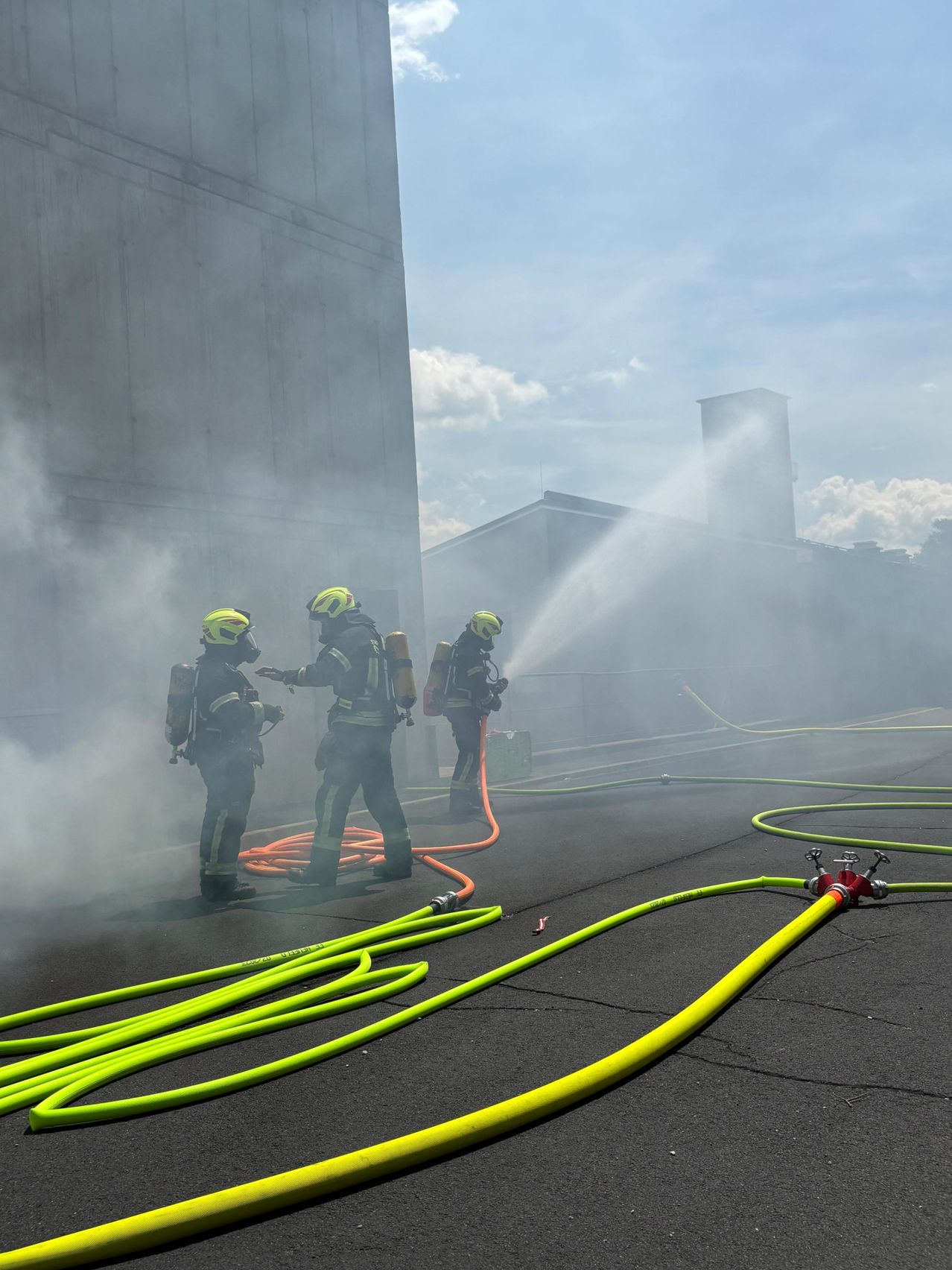 Three firefighters are working on a fire with a hose, one spraying water while two others stand ready with their equipment in a foggy area.
