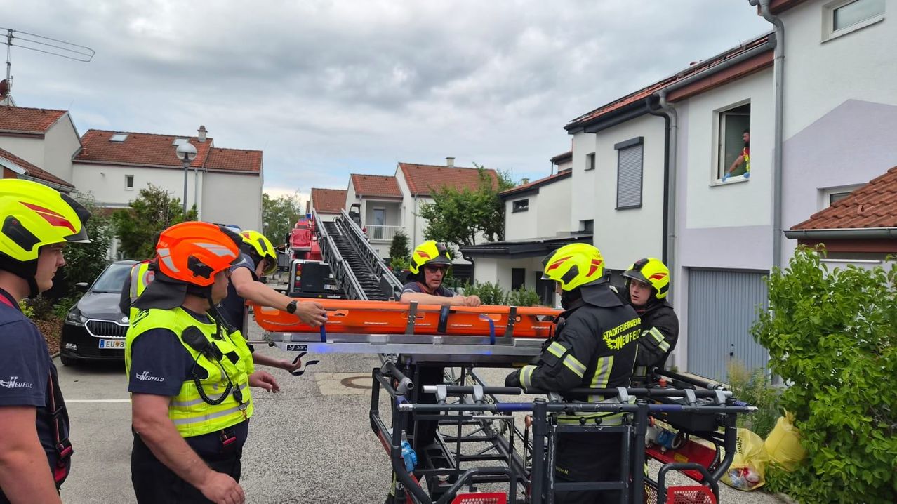 Feuerwehrleute in gelben und schwarzen Uniformen bereiten eine orangefarbene Trage für den Transport vor. Sie stehen vor einem Feuerwehrwagen und einem Gebäude.