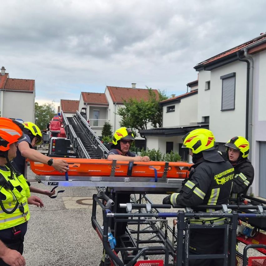 Feuerwehrleute in gelben und schwarzen Uniformen bereiten eine orangefarbene Trage für den Transport vor. Sie stehen vor einem Feuerwehrwagen und einem Gebäude.