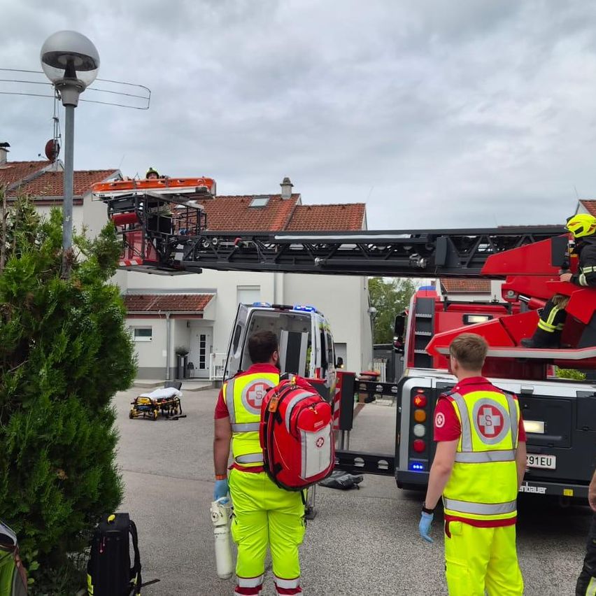 Ein Feuerwehrwagen ist vor einem Haus geparkt. Zwei Personen in gelben Uniformen stehen vor dem Feuerwehrwagen. Eine Person sitzt auf der Leiter des Feuerwehrwagens. Eine weitere Person steht auf der Leiter.