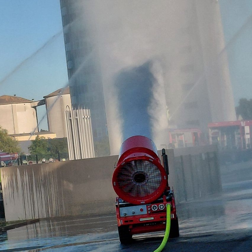Ein rotes Fahrzeug mit einem großen runden Lüfter, das Wasser versprüht, in einem nebligen Bereich, möglicherweise zum Kühlen oder Reinigen. Hohe Gebäude und Bäume sind im Hintergrund.