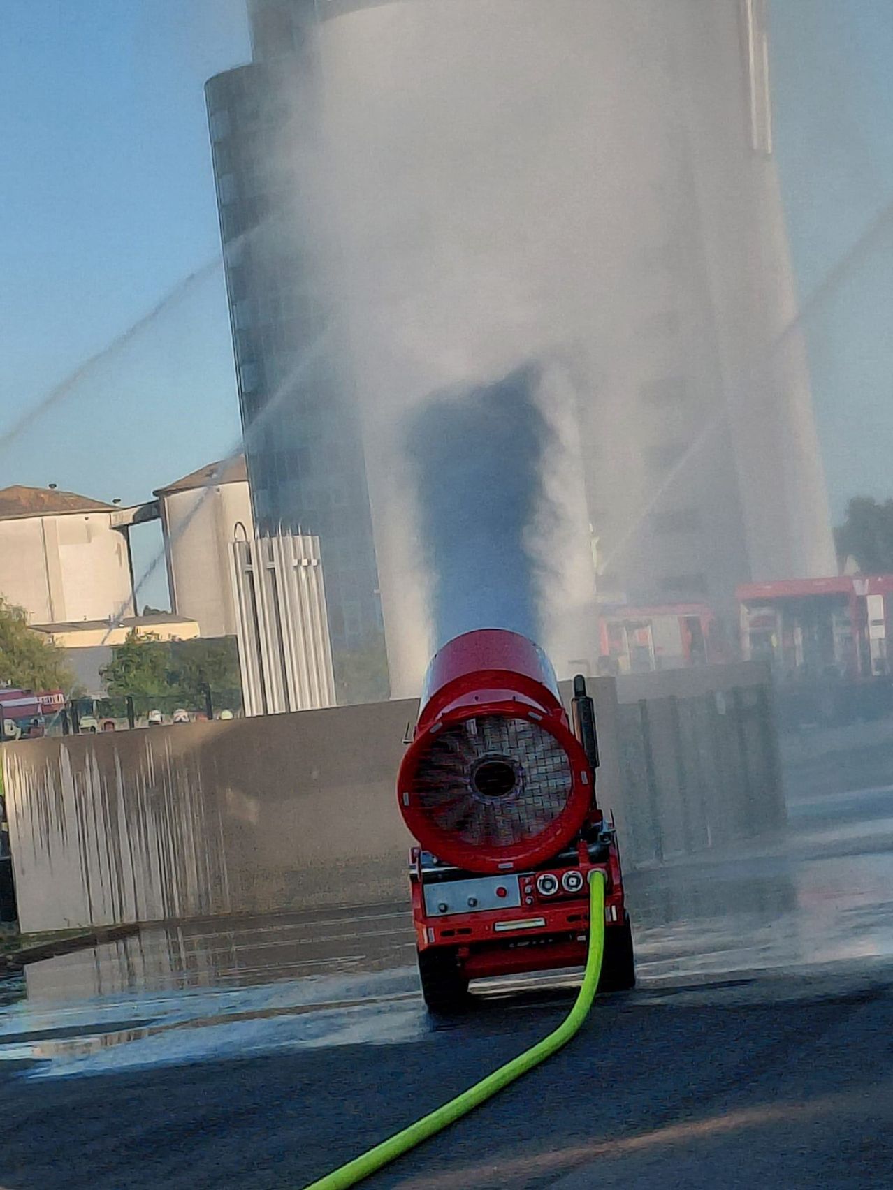 Ein rotes Fahrzeug mit einem großen runden Lüfter, das Wasser versprüht, in einem nebligen Bereich, möglicherweise zum Kühlen oder Reinigen. Hohe Gebäude und Bäume sind im Hintergrund.
