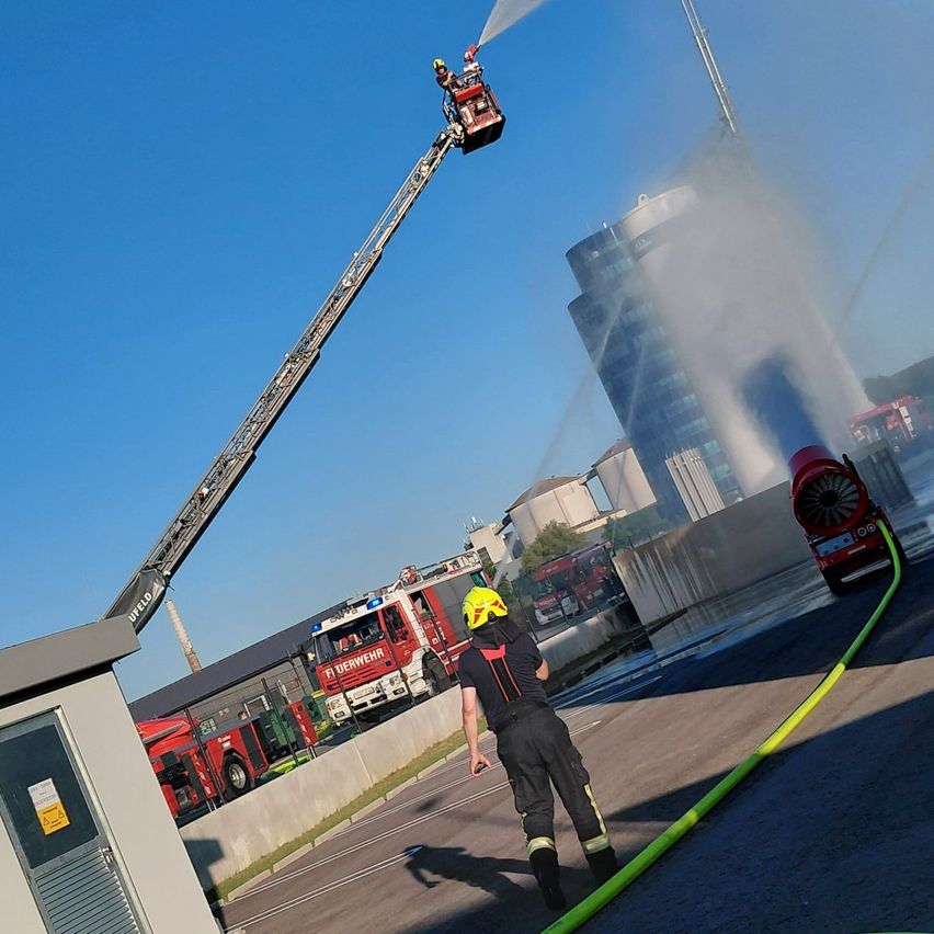 Feuerwehrleute auf einer Leiter spritzen Wasser aus einem Feuerwehrwagen. Ein Gebäude im Hintergrund.