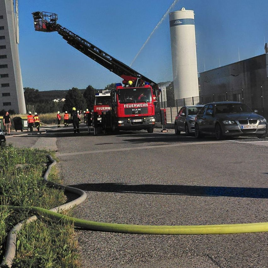 Ein Feuerwehrauto sprüht Wasser auf ein Gebäude mit einem hohen Silo. In der Nähe sind eine Gruppe von Feuerwehrleuten und Autos.