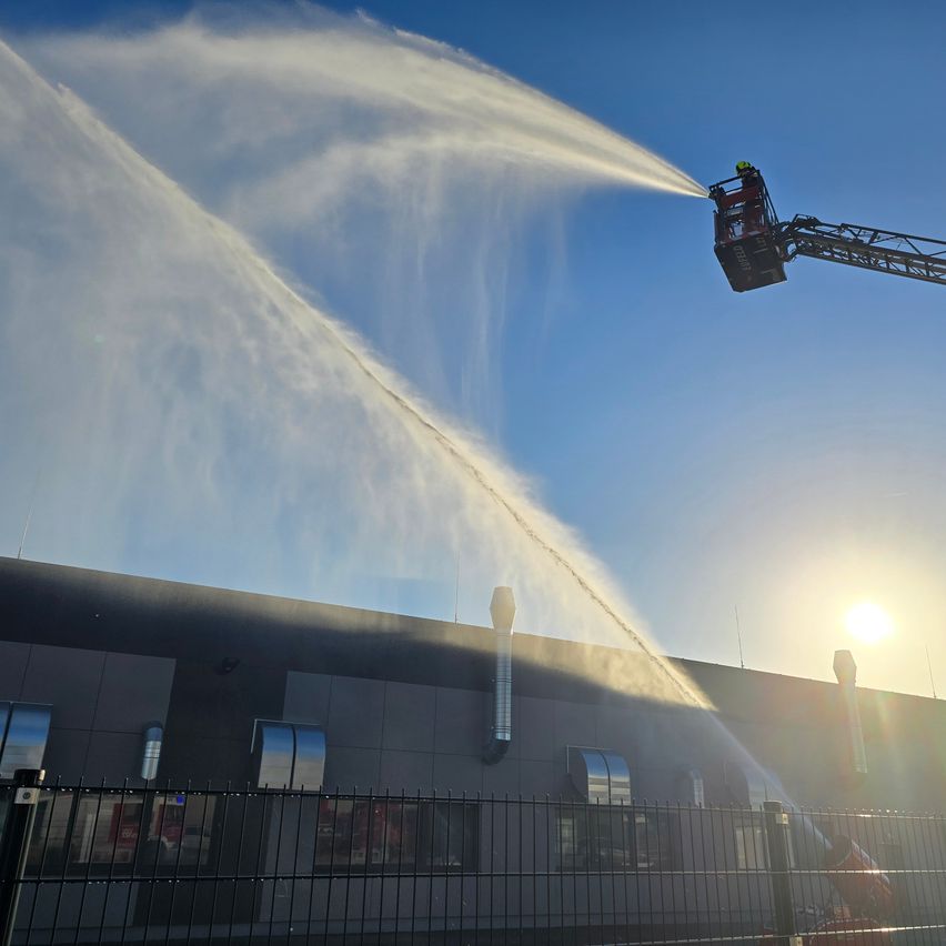 Feuerwehrleute benutzen eine Leiter, um Wasser aus einem Feuerwehrauto auf ein Gebäude bei sonnigem Wetter zu sprühen.